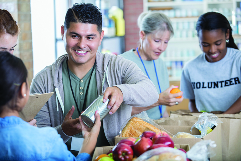 Handsome Hispanic man accepts a canned food donation while volunteering during community food drive. Female volunteers are working in the background.