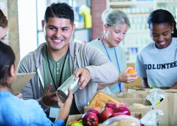 Handsome Hispanic man accepts a canned food donation while volunteering during community food drive. Female volunteers are working in the background.