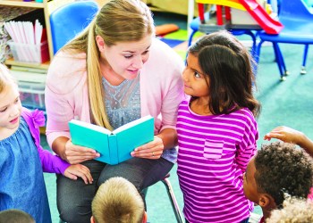 A preschooler teacher in the classroom reading to her class. The multiracial children are sitting on the floor looking up at her. She is talking to a little girl of Pacific Islander ethnicity who is kneeling beside her.