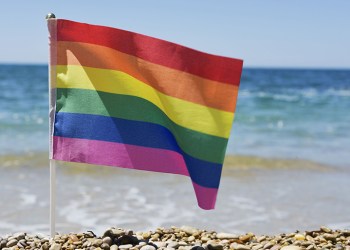 closeup of a small rainbow flag in a pole stuck in a shingle beach, with the ocean in the background