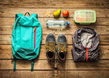 Traveling - packing (preparing) for adventure school trip concept. Backpack, boots, jacket, lunch box, water and fruits on wooden background captured from above (flat lay).