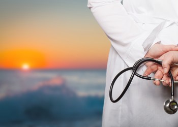 The doctor put his hands with stethoscope behind his back on a blurred background.