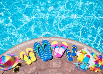 Swimming pool accessories flat lay. Top view of beach items on pool deck. Flip flops, bikini and hat, sun glasses. Water toys. Summer vacation in tropical resort. Copy space. Colorful beach wear.