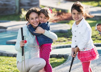 A young family playing miniature golf outdoors on a sunny day. A Hispanic mother with her two daughters, 3 and 4 years old. She is hugging the younger girl, while the older one is swinging a golf club.