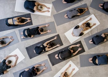 An aerial shot of a multi ethnic group of men and women practice yoga on on mats while wearing grey, black and white in an industrial setting. They are reaching forward in child's pose.