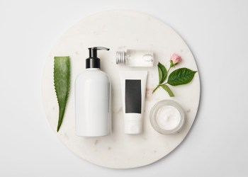 Top view of different cosmetic containers, aloe vera leaf and rose flower on white round surface