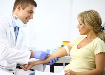 Middle aged blond woman visiting general practitioner.Both are sitting in examination room.The doctor is drawing blood sample from her vein.She has dark blond hair pulled back with a clip.There's a little smir on her face because this is painless procedure.