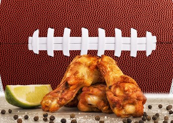 Chicken wings with a lime wedge, salt and peppercorn on concrete table. Football ball image in background.