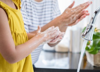 Unrecognizable mom helps her young daughter wash her hands. They are rubbing their hands together creating foam with the soap.