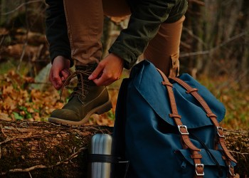 Female hiker tying shoelaces outdoors in autumn forest, near thermos and backpack. View of legs. Hiking and leisure theme