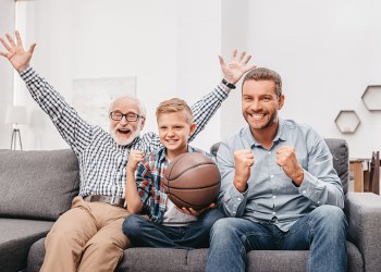 Little boy on couch with grandfather and father, cheering for a basketball game and holding a basketball ball