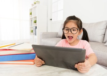 cheerful young female children holding mobile digital tablet sitting in living room and using online e-learning system studying.