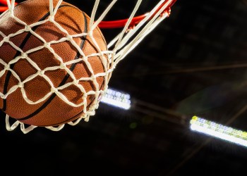 A close-up view looking up at an orange basketball falling through the rim and a white nylon net with the arena lights and lens flare in the background.