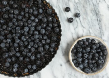 An overhead extreme close up horizontal photograph of a homebred double blueberry tart and a small white bowl of fresh blueberries.