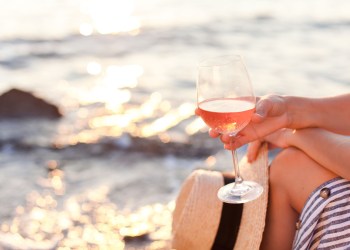 Girl is sitting on sea beach with wineglass of wine at sunset in summer vacation in resort. Tourist woman in striped dress with straw hat is enjoying life, relaxing, drinking, traveling.