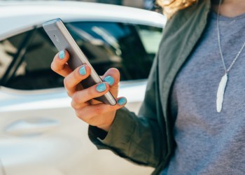 Young woman using smart phone standing on street near road and car. Female's hands holding mobile device in city, close-up.