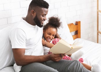 Story time. Loving african american dad reading fairy tales to his little daughter, sitting together on bed