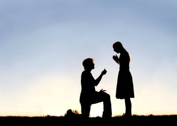 A silhouette of a young man, down on one knee and holding a diamond engagement ring, proposing to his girlfriend for engagement and wedding