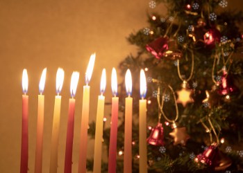 Hanukkah candles burning on the background of Christmas tree, decorated with bells and stars of David. Concept of two holidays happening in the same period this year. Be the Light