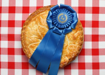 Best of the Best pie with a blue first place ribbon on top. The pie is sitting on a classic red checkerboard tablecloth. Image evokes the feeling of classic Americana. Camera angle is from directly above.