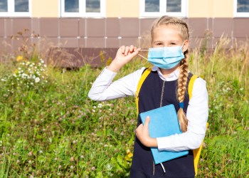 A girl in a blue school uniform stands in front of the school building in a medical mask, holds a textbook in her hands, looks at the camera. Back to school. Security concept, virus protection, new normal.