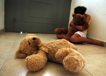 A young girl who is a victim of domestic violence and sexual abuse sits on the floor next to the front door and gets comfort from her teddy bears