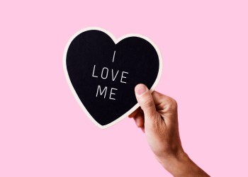 closeup of a man holding a black heart-shaped sign with the text I love me written in it, on a pink background -love yourself