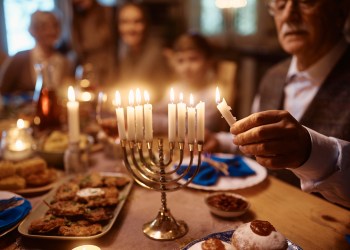 Close up of extended Jewish family celebrating Hanukkah Chanukah at dining table. Focus is on mature man lighting candles in menorah.