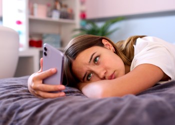 Depressed Teenage Girl Lying On Bed At Home Looking At Mobile Phone - kids in crisis