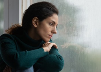 Thoughtful stressed young hispanic latin woman sitting on windowsill, looking outside on rainy weather, having depressive or melancholic mood, suffering from negative thoughts alone at home. loneliness