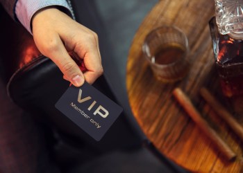 Man holds VIP member card. View from the top on the gentleman's hand that holds exclusive VIP membership card next to the wooden table with whisky in carafe and glass with cigars.