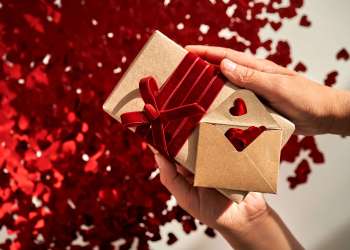 A craft gift and an envelope with many red shiny hearts on a background in the hands. A wonderful photo for Valentine’s Day.