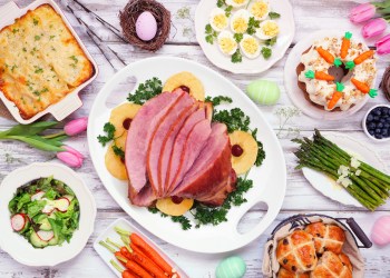 Traditional Easter ham dinner. Top view table scene on a white wood background. Ham, scalloped potatoes, eggs, hot cross buns, carrot cake and vegetables.